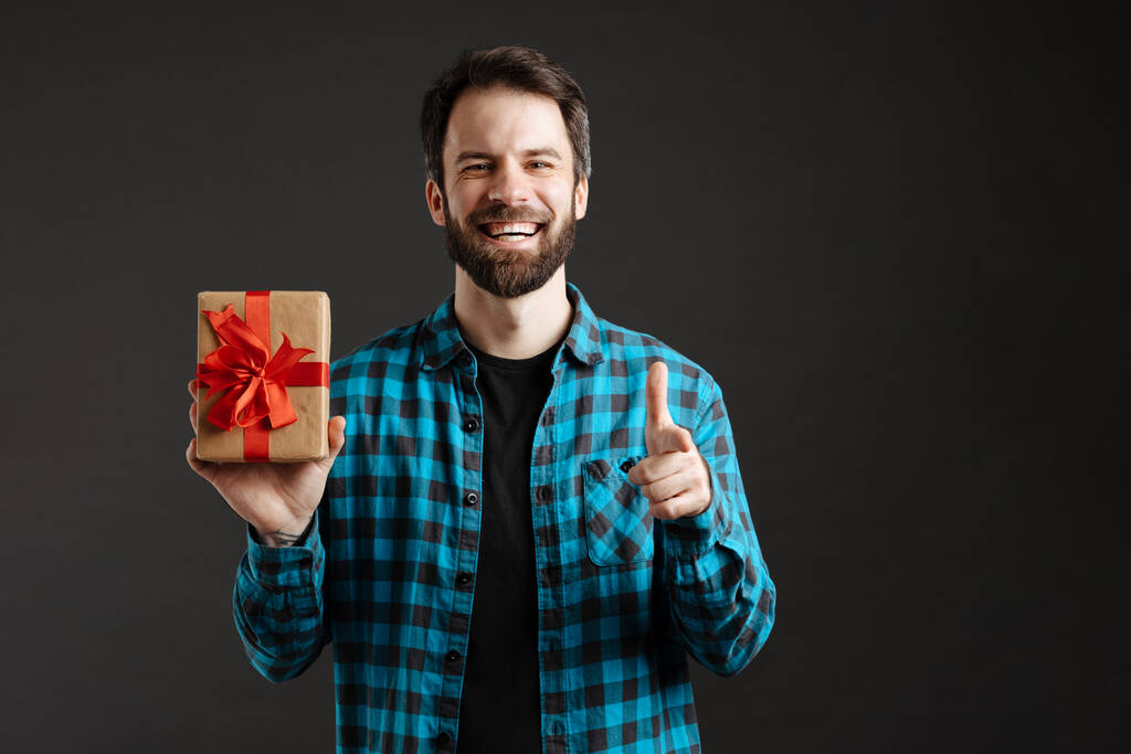 baard gelukkig man wijzend vinger naar camera terwijl het tonen van geschenk doos geïsoleerd over zwarte achtergrond - Foto, afbeelding