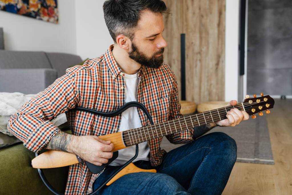 Mediados de edad morena barbudo hombre tocando la guitarra acústica mientras está sentado en un piso en la sala de estar - Foto, Imagen