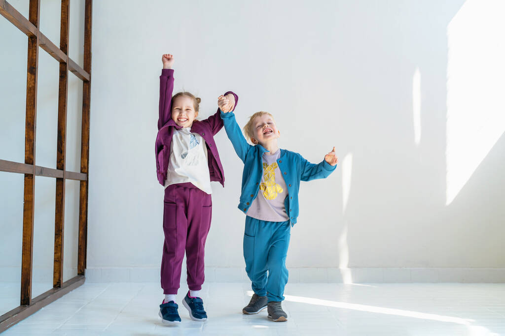 Malý šťastný chlapec a dívka v módním oblečení úsměv, póza a skok na pozadí bílé stěny studia. koncept reklamy na dětské oblečení, emocionální děti, dynamický image - Fotografie, Obrázek