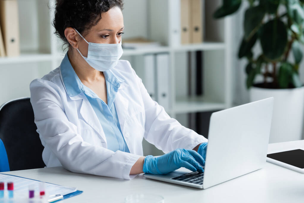 Scientifique afro-américain en masque médical à l'aide d'un ordinateur portable près d'une tablette numérique et des éprouvettes floues  - Photo, image