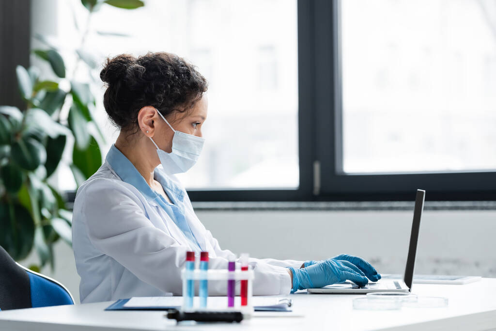 Vista lateral do cientista afro-americano em máscara médica e luvas de látex usando laptop perto de tubos de ensaio desfocados  - Foto, Imagem