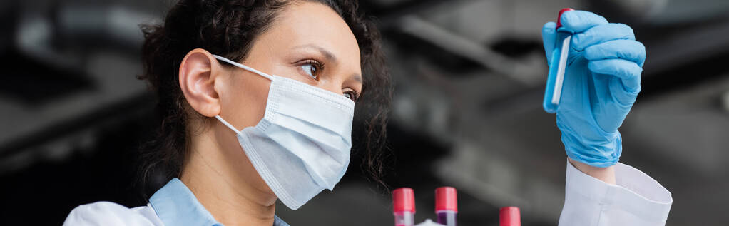 Χαμηλή γωνία άποψη του Αφροαμερικανού επιστήμονα στην ιατρική μάσκα κοιτάζοντας δοκιμαστικό σωλήνα, πανό  - Φωτογραφία, εικόνα