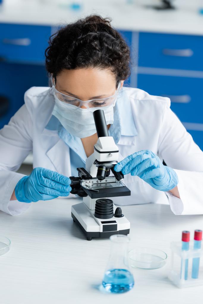 Cientista afro-americano em óculos de segurança e máscara médica trabalhando com microscópio perto de tubos de ensaio  - Foto, Imagem