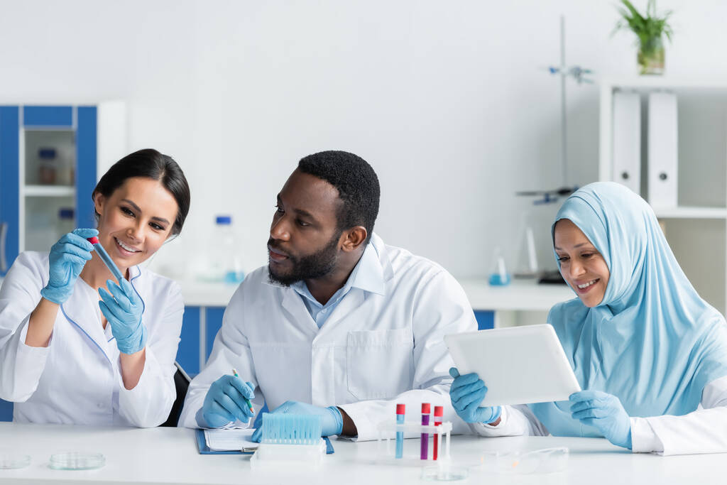 Smiling scientist holding test tube near interracial colleagues with clipboard and digital tablet  - Foto, Imagen