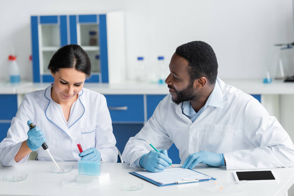 Smiling african american scientist in latex gloves writing on clipboard and looking at colleague with test tube  - Photo, Image