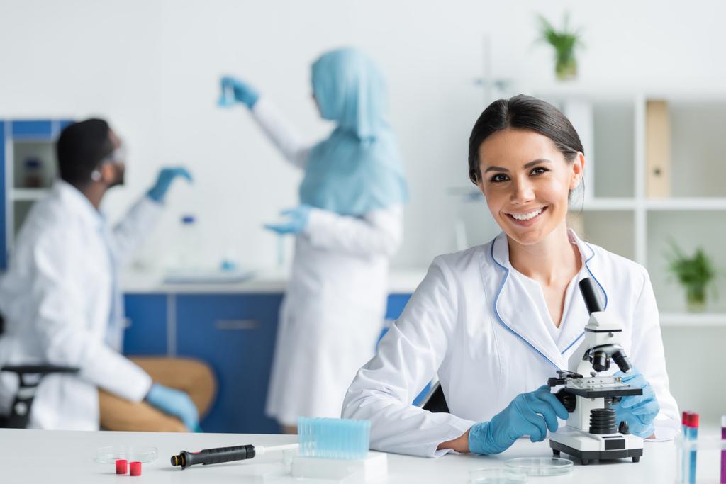 счастливый ученый в латексных перчатках улыбается в камеру возле микроскопа и пробирки  - Фото, изображение