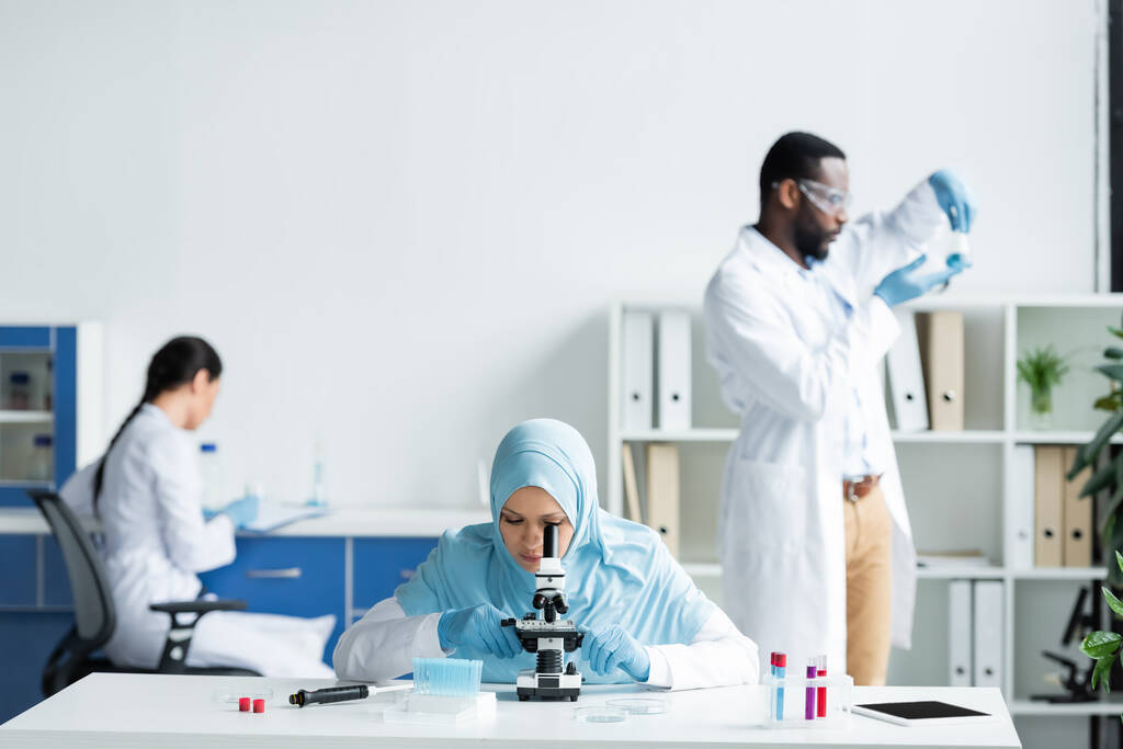 Scientifique musulman regardant au microscope près de tablette numérique et des collègues interraciaux sur fond flou  - Photo, image