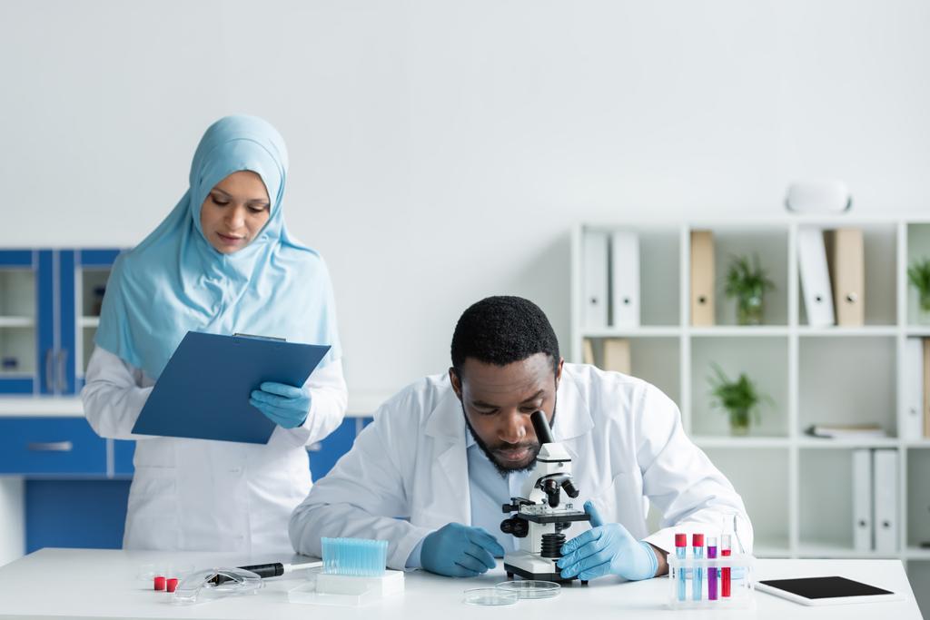 Afrikanischer Wissenschaftler schaut mit Klemmbrett durch Mikroskop neben muslimischem Kollegen  - Foto, Bild
