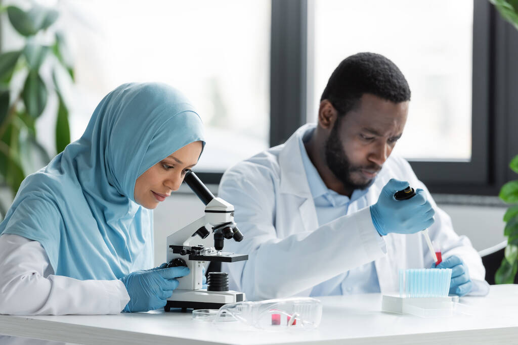 研究室で働くアフリカ系アメリカ人の同僚に近い顕微鏡を通して見るイスラム教徒の科学者  - 写真・画像