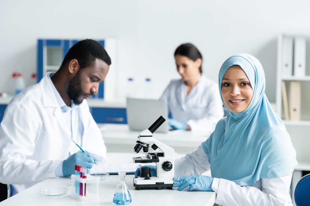 Glimlachende Arabische wetenschapper in hijab kijkend naar camera in de buurt van microscoop en Afrikaanse Amerikaanse collega in laboratorium  - Foto, afbeelding