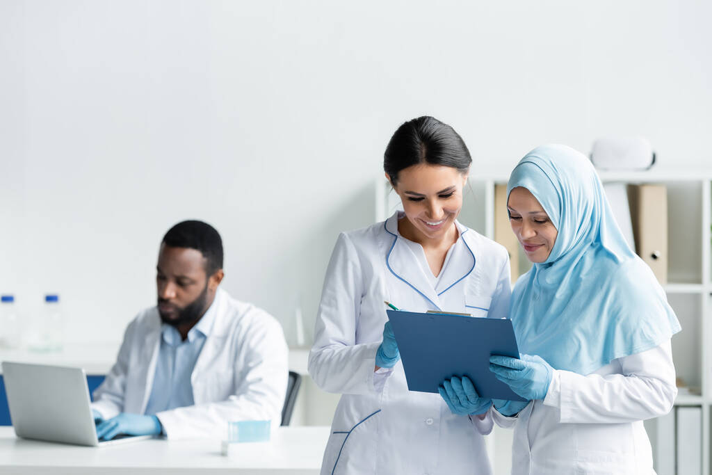 межрасовые ученые улыбаются, держа планшет рядом с размытым африканским американским коллегой с ноутбуком в лаборатории  - Фото, изображение