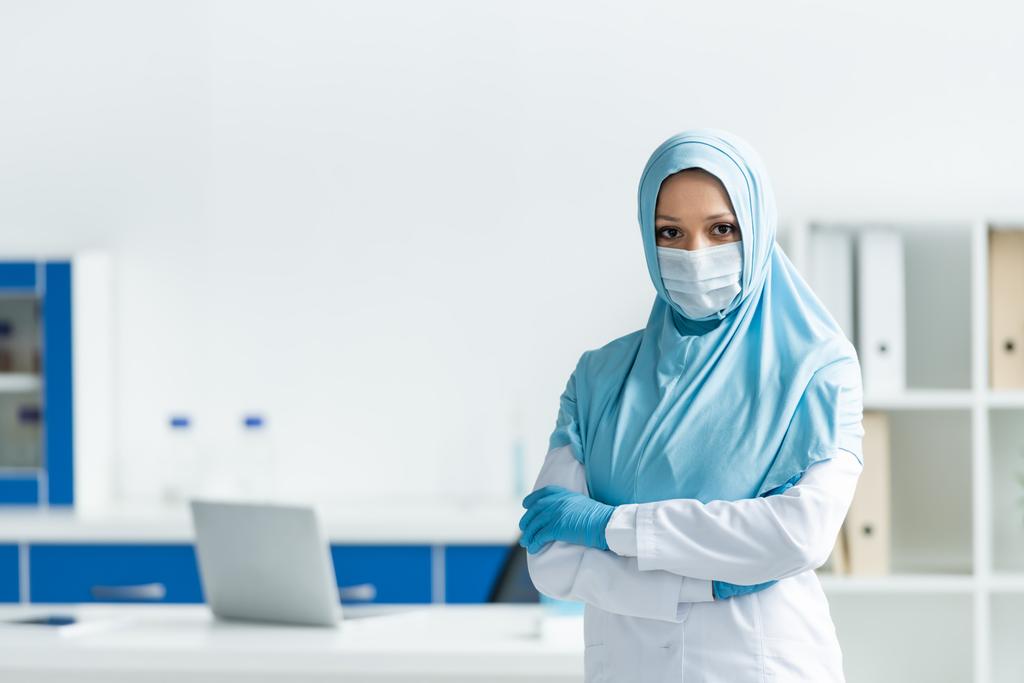 Tıbbi maskeli ve tesettürlü Müslüman doktor klinikte çapraz kollarla ayakta duruyor  - Fotoğraf, Görsel