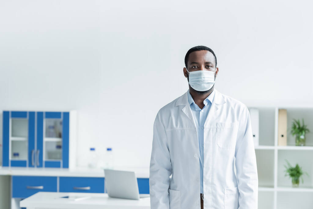 Tıbbi maskeli Afrikalı Amerikalı bilim adamı ve kameraya bakan beyaz önlüklü.  - Fotoğraf, Görsel