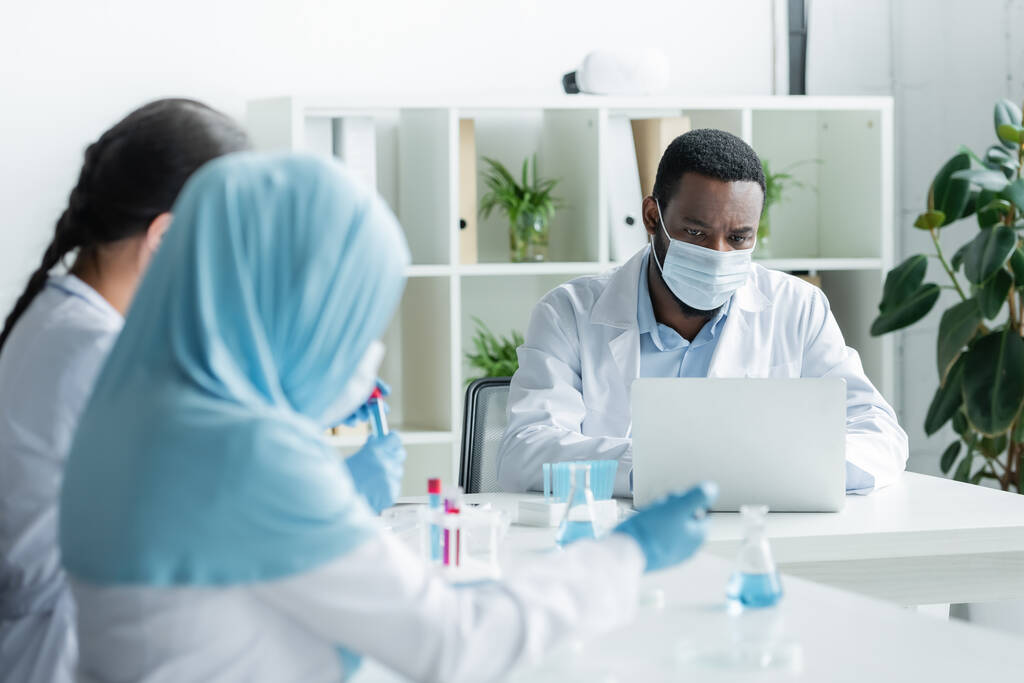 Африканський американський вчений медичної маски за допомогою ноутбука біля розмитих колег з обладнанням. - Фото, зображення