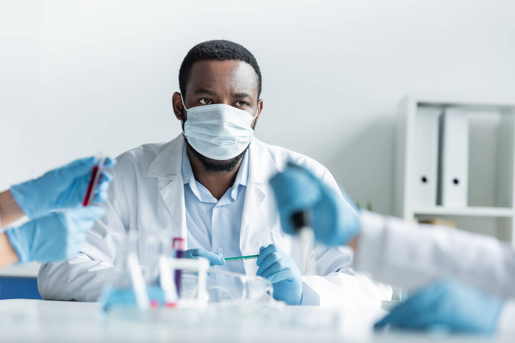 Αφροαμερικανός επιστήμονας με προστατευτική μάσκα κοιτάζοντας θολή συναδέλφους που εργάζονται στο εργαστήριο  - Φωτογραφία, εικόνα