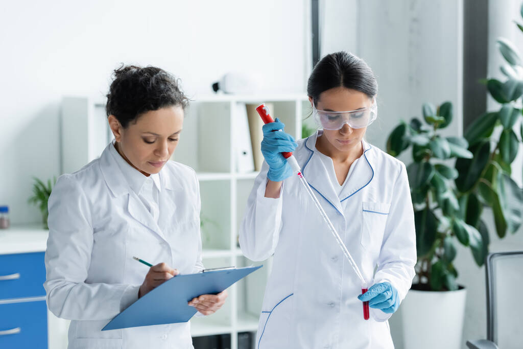 African american scientist writing on clipboard near colleague with test tube and pipette  - Foto, imagen