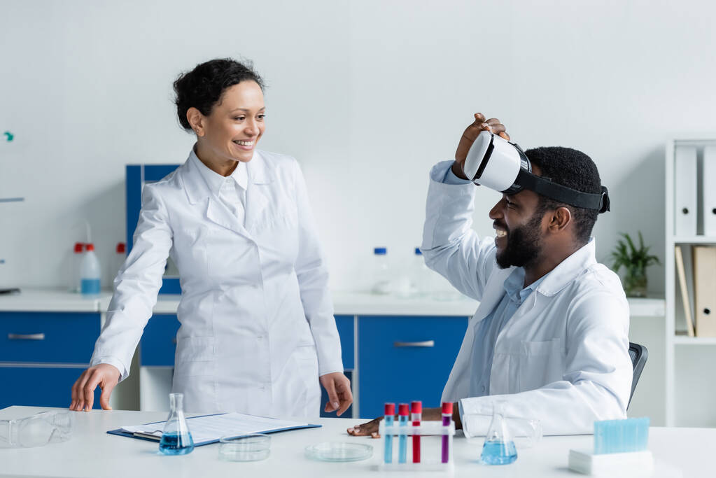 Африканский американский ученый, стоящий рядом с коллегой в наушниках рядом с оборудованием в лаборатории - Фото, изображение