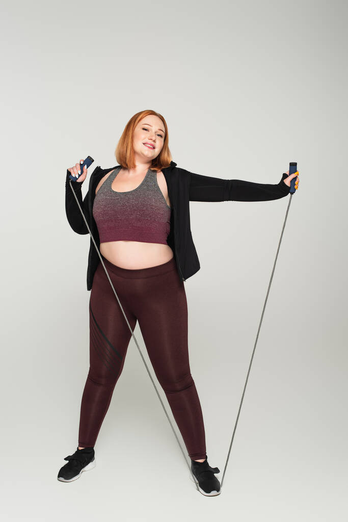 Smiling plus size sportswoman holding corda da salto su sfondo grigio - Foto, immagini