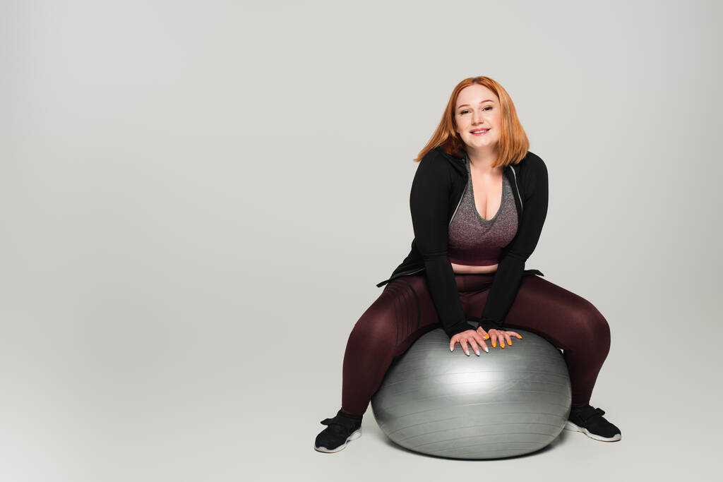 Vrolijke plus size sportvrouw zittend op fitness bal op grijze achtergrond - Foto, afbeelding
