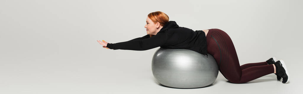 Вид сбоку плюс размер женщины, тренирующейся на фитнес-мяче на сером фоне, баннер  - Фото, изображение