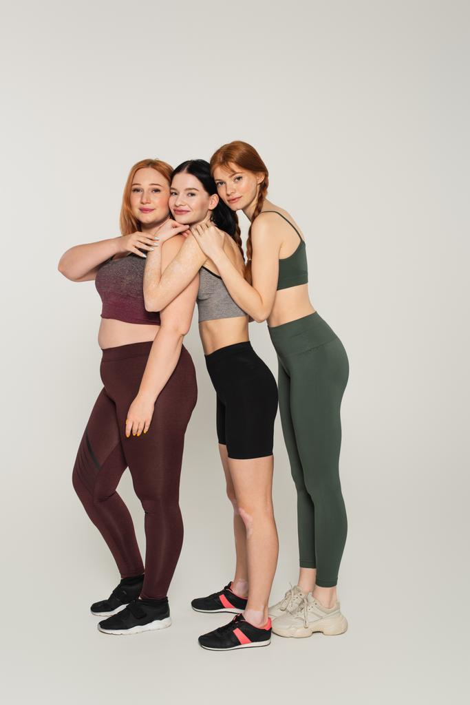 Full length of smiling body positive women in sportswear embracing each other on grey background - Photo, Image