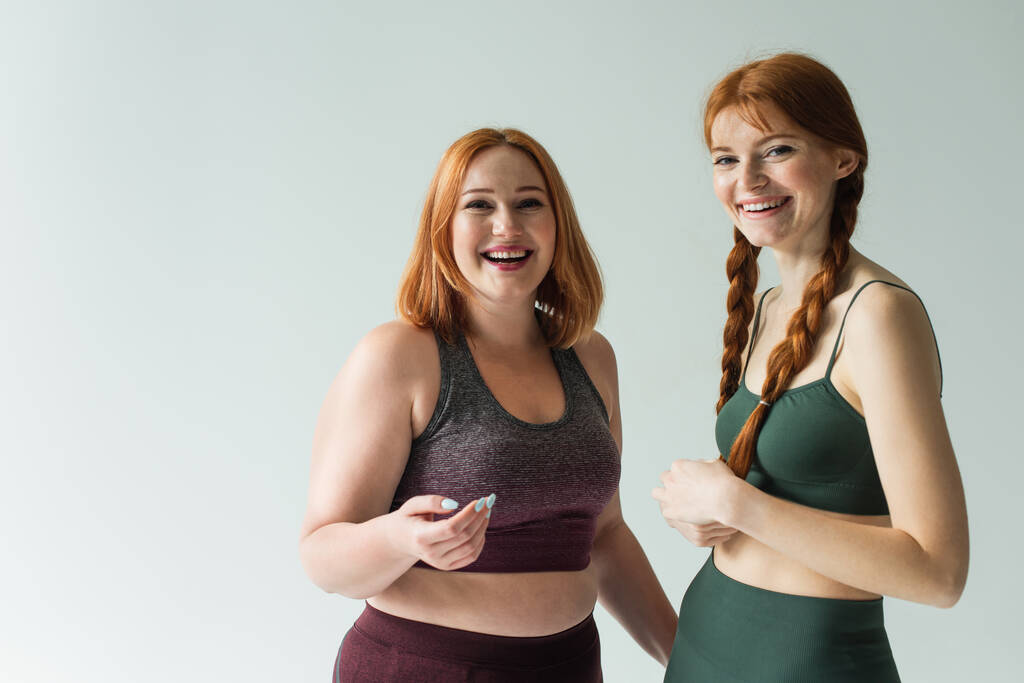 Smiling body positive sportswomen looking at camera isolated on grey  - Photo, Image