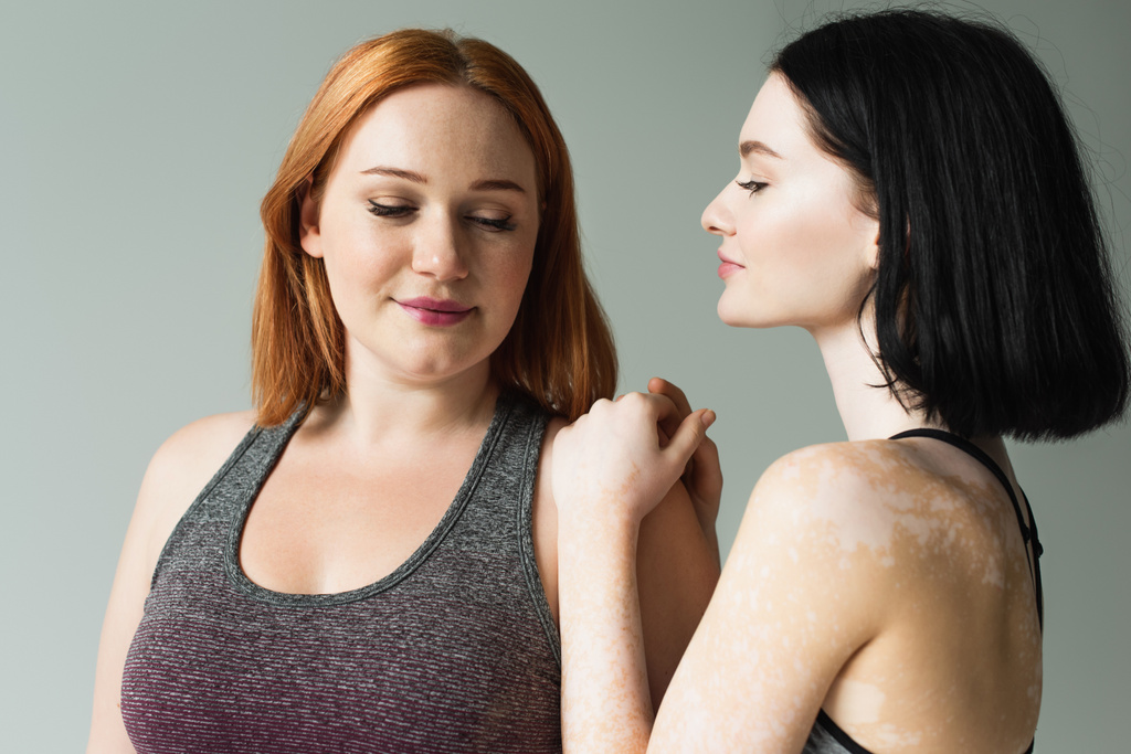 Sportvrouw met vitiligo op zoek naar plus size vriend geïsoleerd op grijs  - Foto, afbeelding