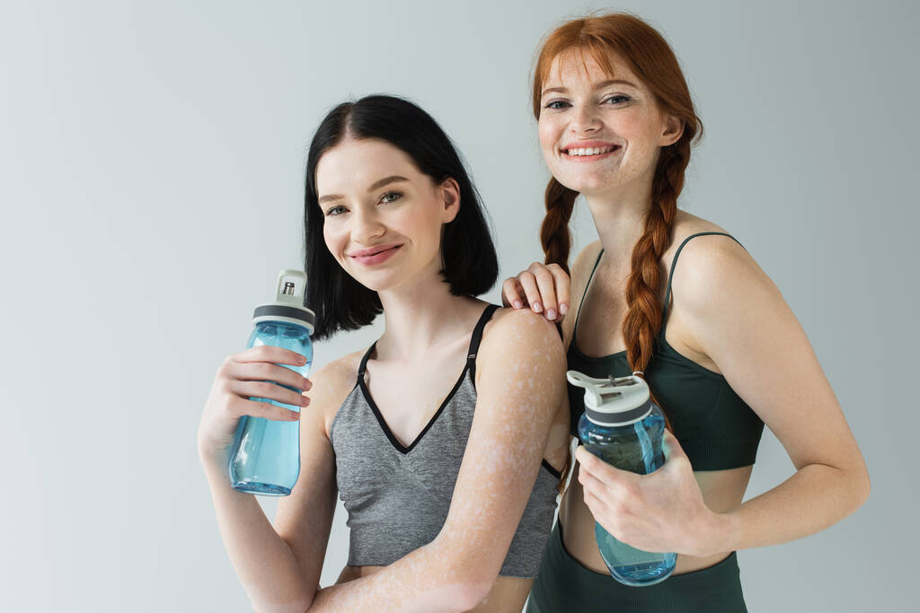 Cheerful sportswomen with vitiligo and freckles holding sports bottles isolated on grey  - Photo, Image