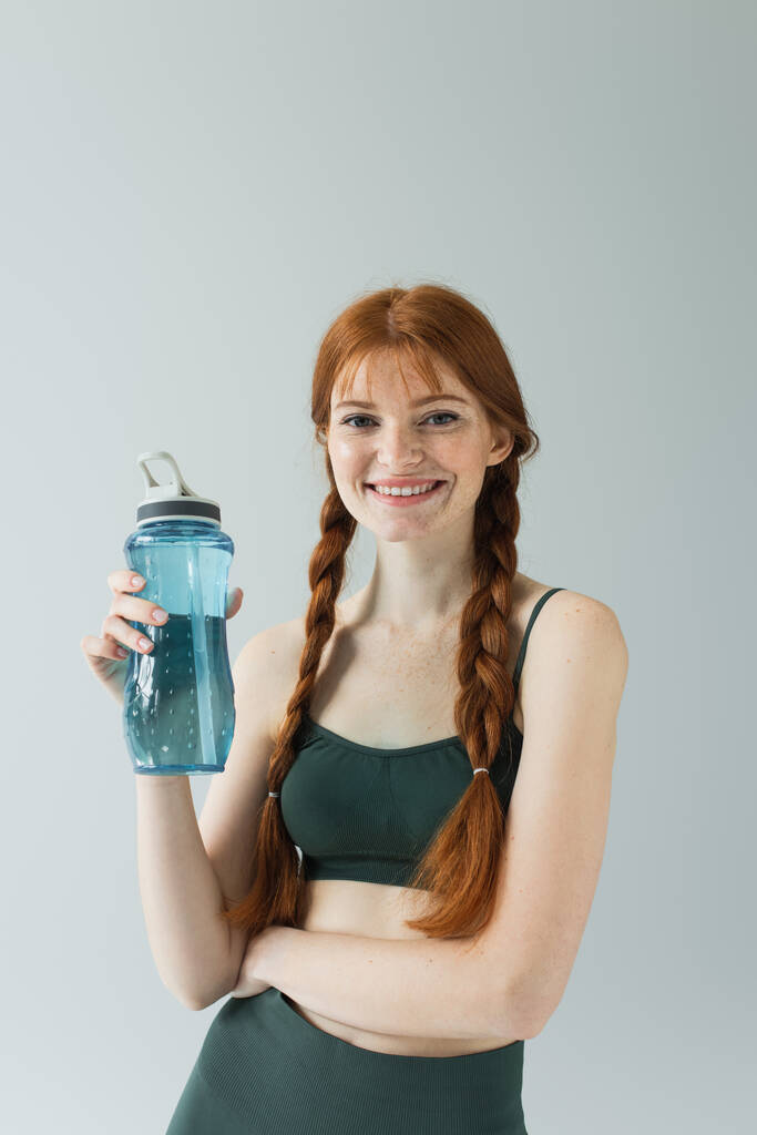 Felice donna sportiva lentigginosa che tiene bottiglia sportiva isolata su grigio  - Foto, immagini