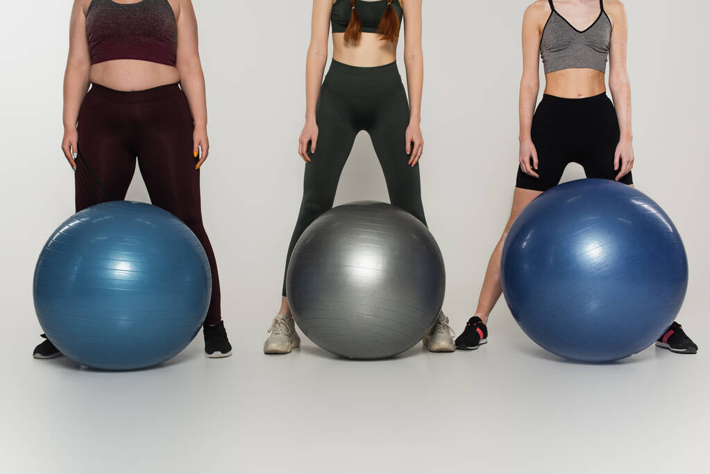 Gesneden weergave van het lichaam positieve sportenvrouwen staan in de buurt van fitness ballen op grijze achtergrond - Foto, afbeelding
