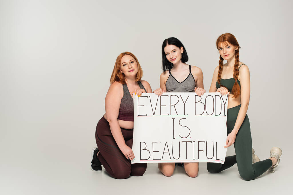 Body positive women in sportswear holding placard with every body is beautiful lettering on grey background - Photo, Image