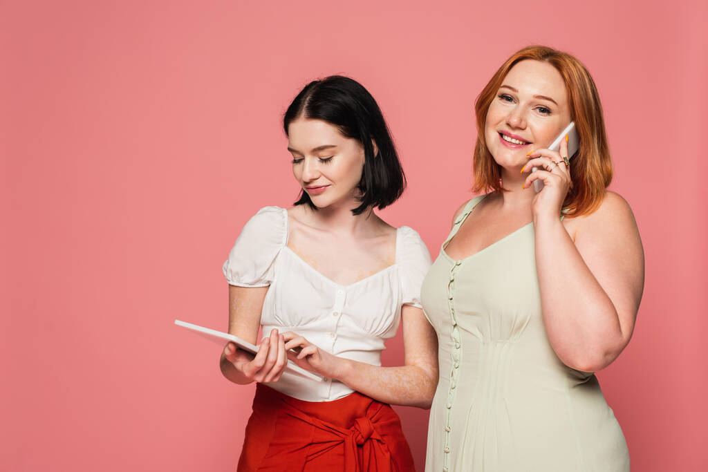Hymyilevä plus koko nainen puhuu älypuhelimella lähellä ystävä vitiligo käyttäen digitaalinen tabletti eristetty vaaleanpunainen  - Valokuva, kuva