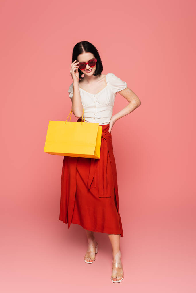 Mujer bonita con vitiligo sosteniendo gafas de sol y bolso de compras sobre fondo rosa  - Foto, Imagen
