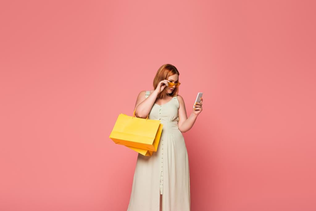 Stylowe ciało pozytywne kobieta gospodarstwa torby na zakupy i smartfon odizolowany na różowy  - Zdjęcie, obraz