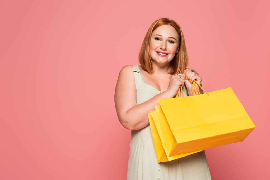 Uśmiechnięta rudowłosa kobieta z torbami na zakupy patrząca na kamerę odizolowana na różowo  - Zdjęcie, obraz