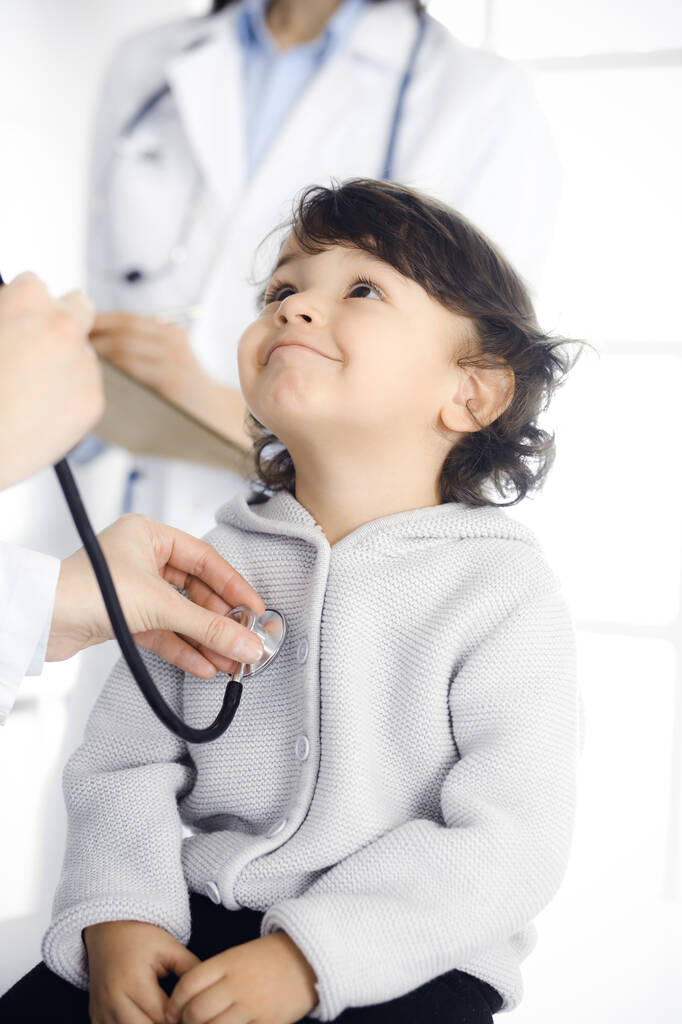 Woman-doctor examining a child patient by stethoscope. Cute arab toddler at physician appointment. Medicine concept - Photo, Image