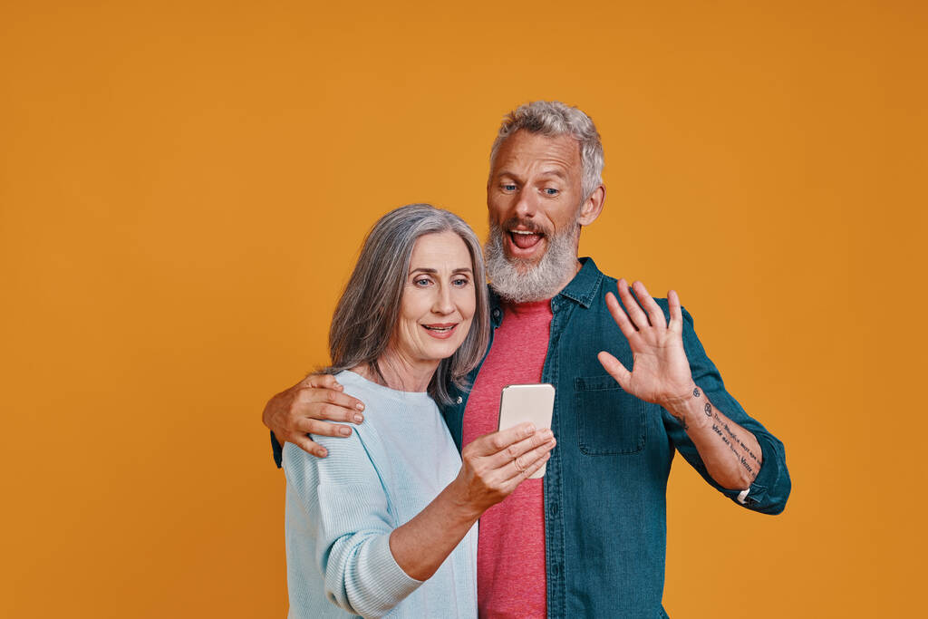 Feliz pareja de ancianos sonriendo y usando teléfonos inteligentes mientras están de pie sobre fondo naranja - Foto, imagen