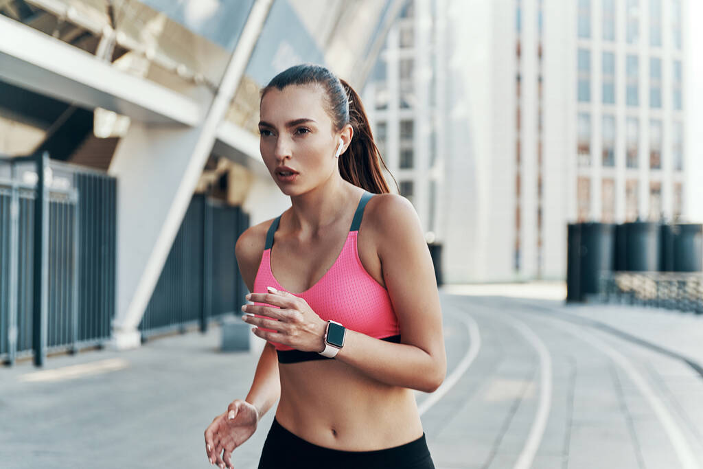 Красива молода жінка в спортивному одязі носить навушники і розумний годинник під час роботи на відкритому повітрі
 - Фото, зображення