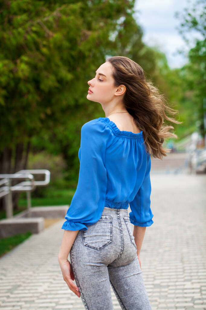 Young beautiful brunette girl in a blue blouse and gray jeans, summer park outdoor  - Photo, Image