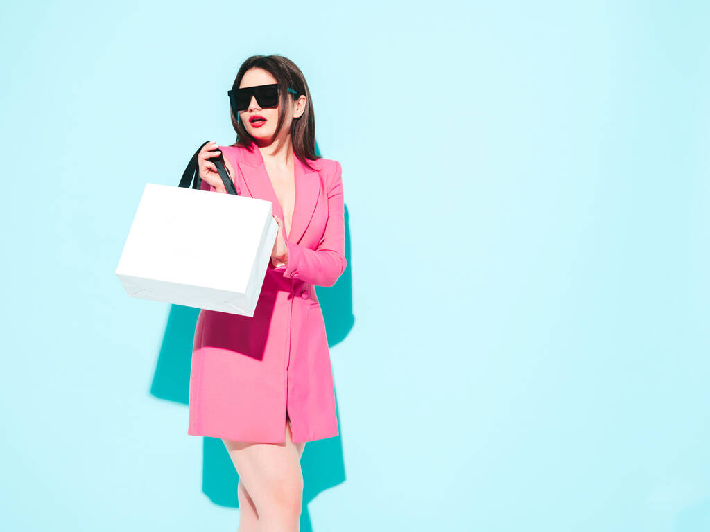 High-Fashion-Porträt der jungen schönen brünetten Frau trägt schöne rosa Sommeranzug. Sexy trendiges Modell posiert in der Nähe der blauen Wand im Studio.Modische Frau mit Sonnenbrille. Die Einkaufstasche halten. - Foto, Bild