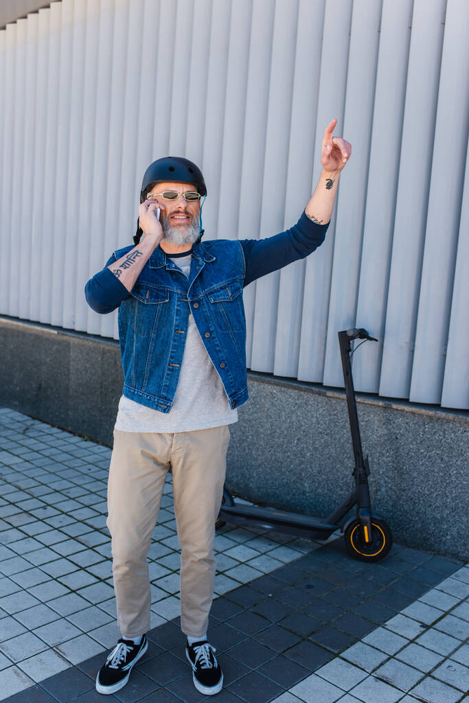 full length of cheerful and mature hipster in helmet and sunglasses talking on smartphone and gesturing near e-scooter  - Photo, Image