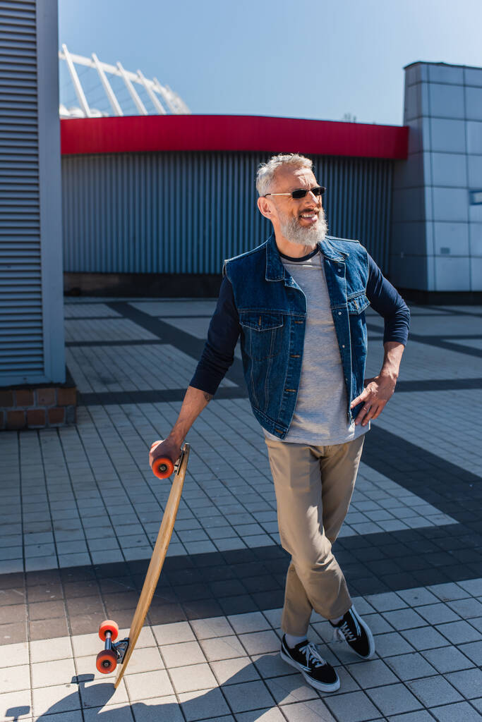 повнометражний щасливий чоловік середнього віку в сонцезахисних окулярах тримає дошку і стоїть з схрещеними ногами на міській вулиці
 - Фото, зображення