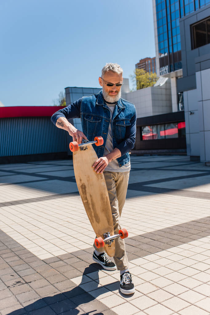 tattooed and middle aged man in sunglasses holding longboard on urban street - Zdjęcie, obraz