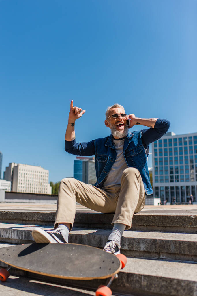 excited middle aged man in sunglasses sitting on stairs near longboard while talking on cellphone and showing rock sign on urban street - 写真・画像