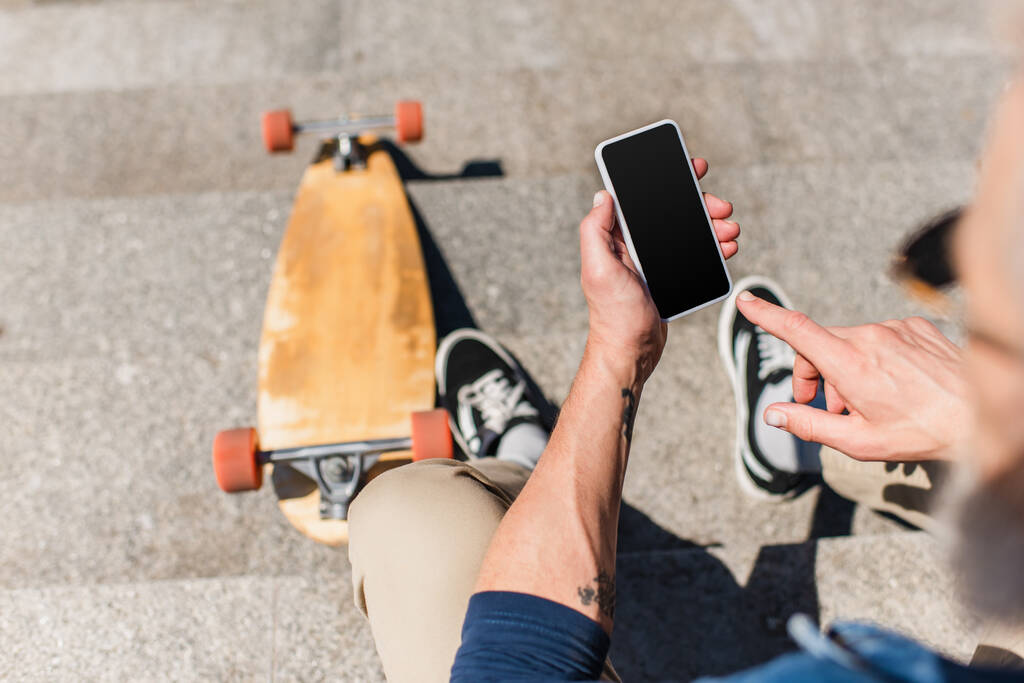top view of tattooed mature man using smartphone with blank screen near longboard on stairs  - 写真・画像