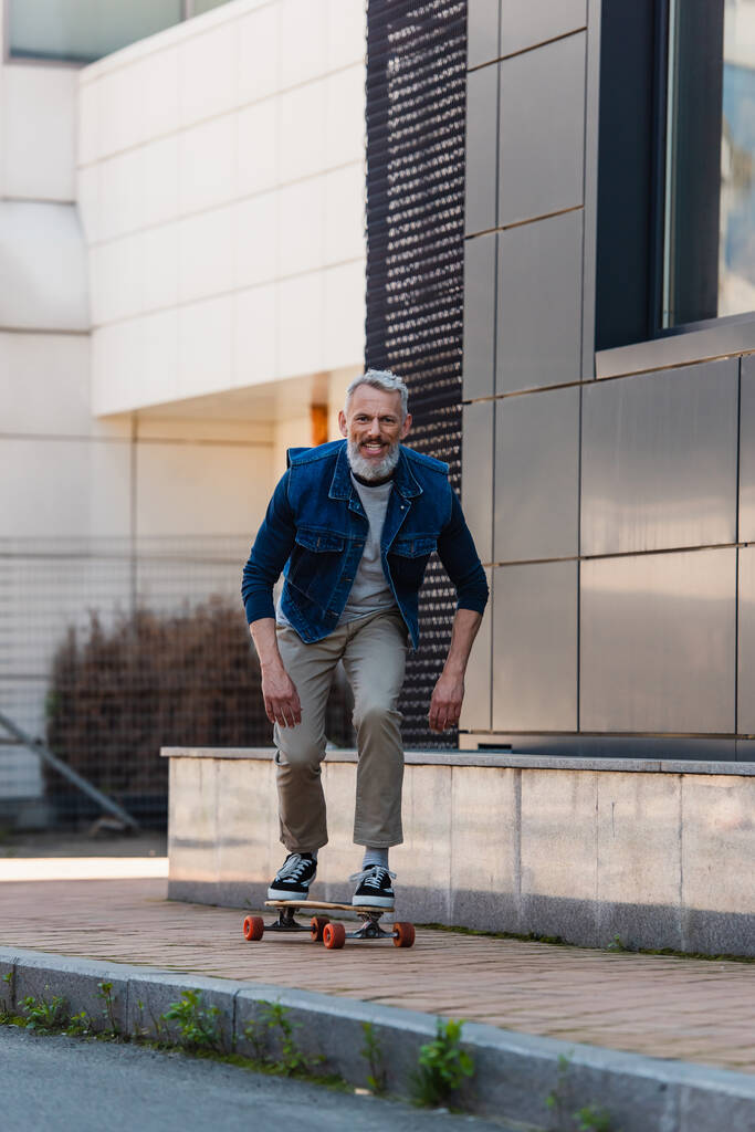 full length of middle aged man smiling and riding longboard on urban street - Foto, immagini