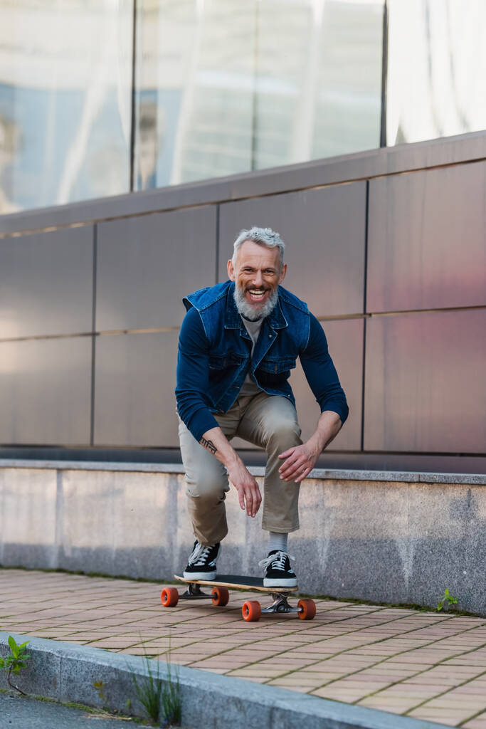 full length of cheerful and middle aged man riding longboard on urban street - Photo, Image