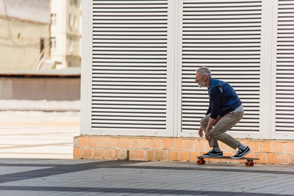 Seitenansicht eines positiven Mannes mittleren Alters, der auf einer städtischen Straße Longboard fährt - Foto, Bild