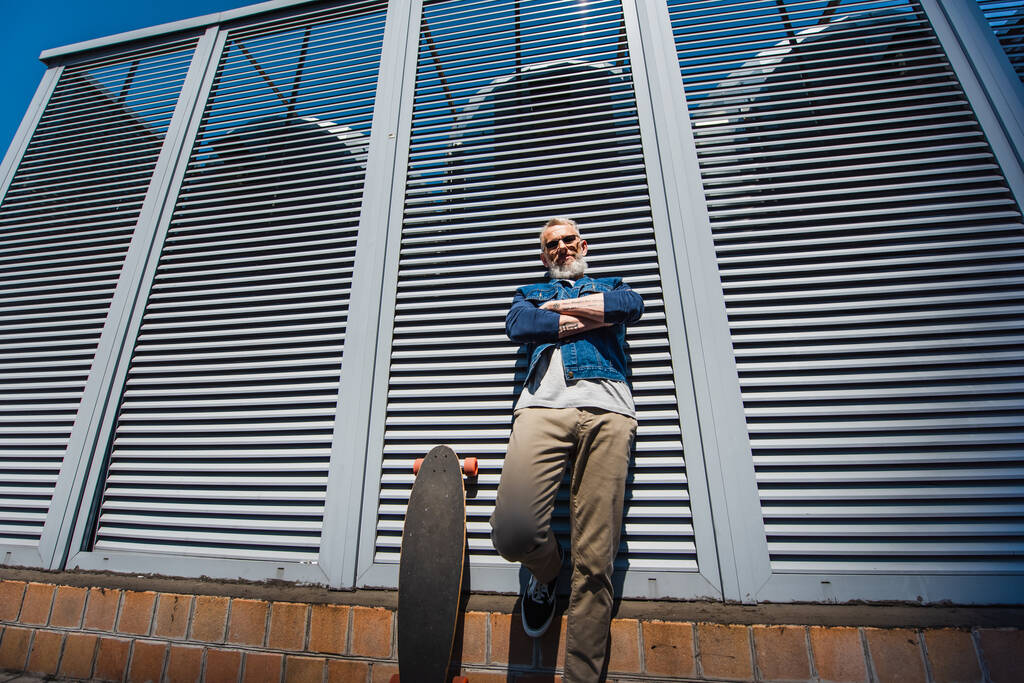 low angle view of positive and middle aged man standing with crossed arms near longboard on street - Photo, image