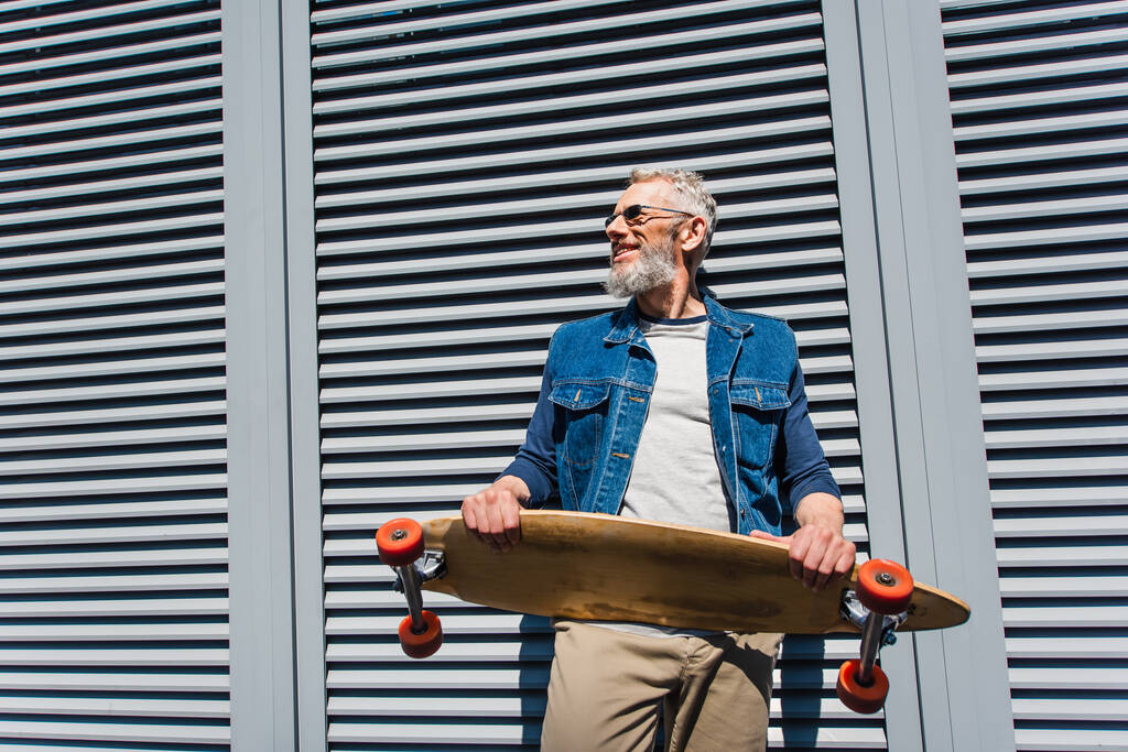 positive and middle aged man in sunglasses holding longboard  - Foto, imagen